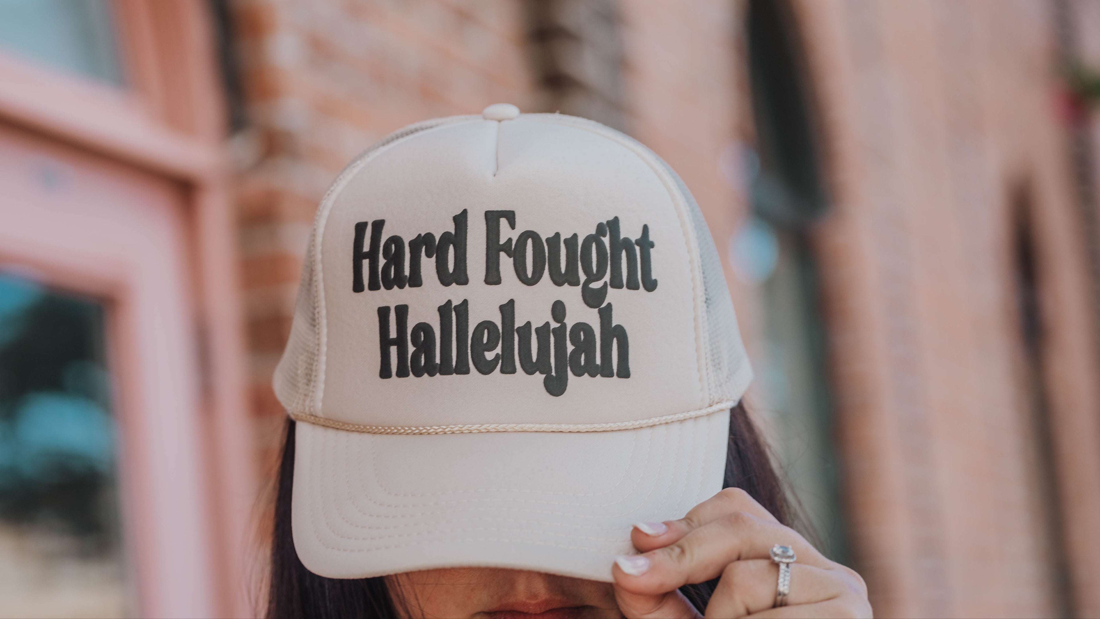 Beige cap with 'Hard Fought Hallelujah' text worn by a person, blurred background